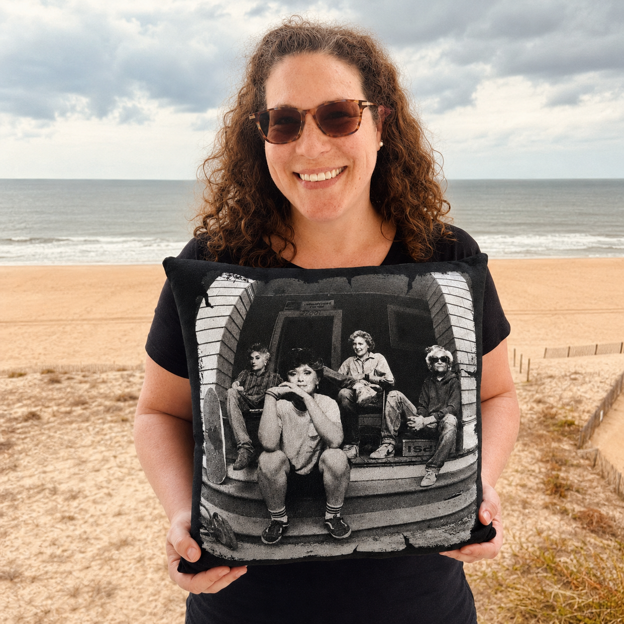 Smiling woman wearing sunglasses at the beach holding a black memory pillow made from a vintage Golden Girls graphic T-shirt.
